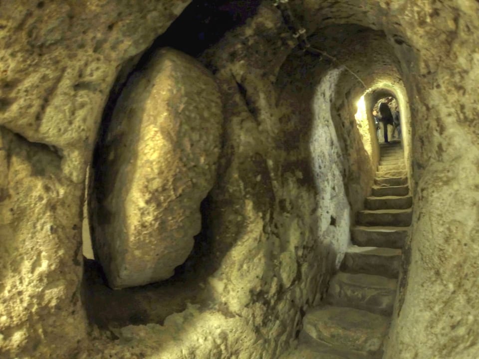 A narrow underground passageway carved out of stone. In the foreground, a large, circular rock that can seal off the entrance has been rolled out of the way.
